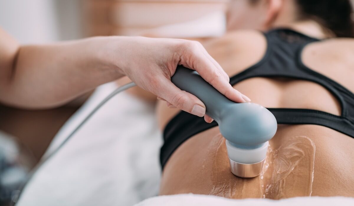 Médico aplica un cabezal de radiofrecuencia sobre la espalda baja de un paciente que está tumbado bocabajo. Se aprecia gel conductor sobre la piel para facilitar la transmisión de la energía térmica. El tratamiento está enfocado en aliviar el dolor lumbar mediante la modulación nerviosa en la clínica Fermaz.