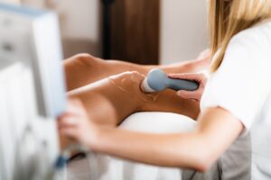 Médico de Fermaz Salud Madrid utilizando un dispositivo de radiofrecuencia sobre la rodilla de un paciente. La rodilla está cubierta con gel conductor. A la izquierda se observa parte del equipo de electromedicina que controla los parámetros del tratamiento intervencionista para el dolor articular crónico.